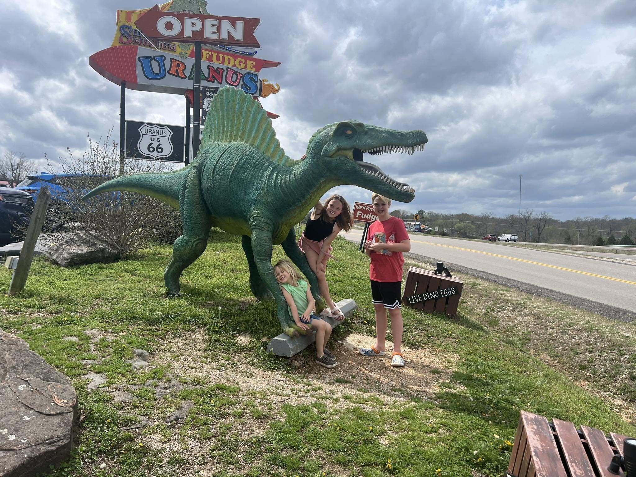 Kids posing with a large green dinosaur statue next to the Uranus US 66 highway shield sign along Route 66 in Missouri