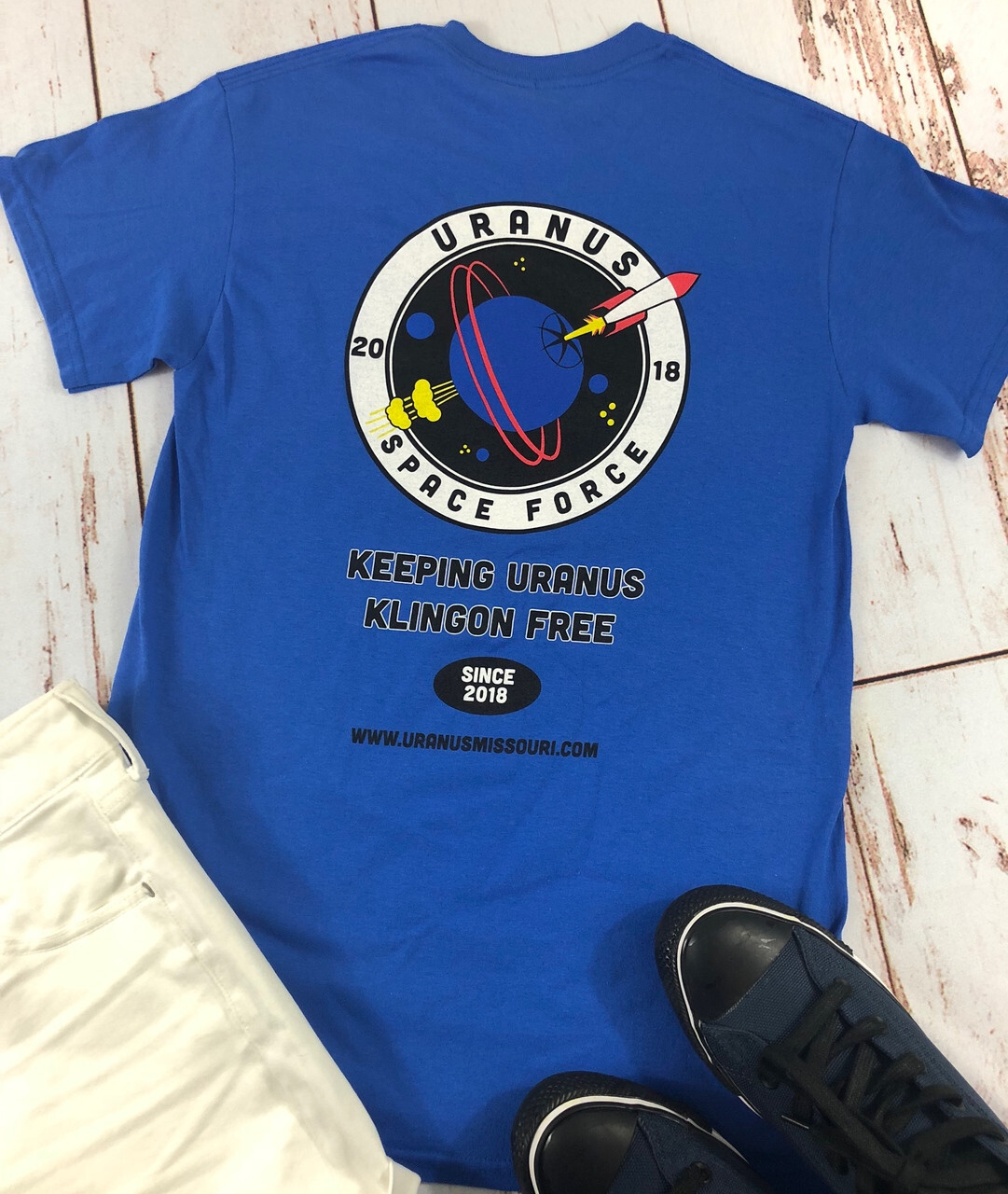 A blue t-shirt with a humorous Uranus Space Force graphic laid flat next to white pants and shoes