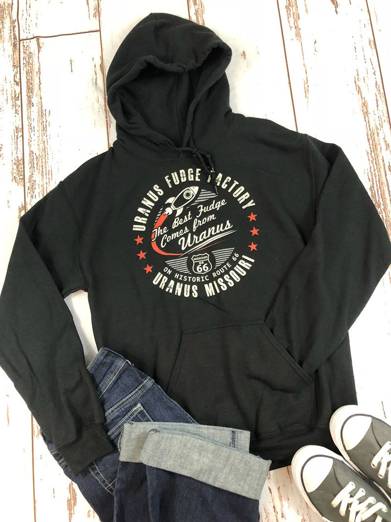 A black hoodie with a Uranus Fudge Factory graphic laid flat with blue jeans and canvas sneakers