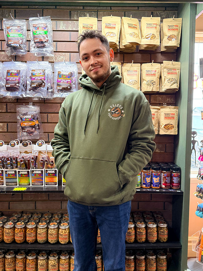 A man standing in a shop wearing an olive green hoodie with a small Uranus Crop Dusters chest logo